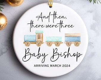 May include: A white ceramic ornament with a blue and brown toy train illustration. The text reads "And then, there were three Baby Bishop Arriving March 2024".