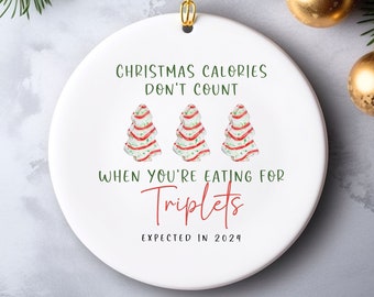 May include: White ceramic Christmas ornament with the text "Christmas Calories Don't Count When You're Eating For Triplets Expected in 2024". Three stylized Christmas trees are depicted with red and white stripes.