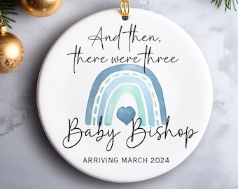 May include: White ceramic Christmas ornament with a blue and green watercolor rainbow, a heart, and the text "And then, there were three. Baby Bishop Arriving March 2024."