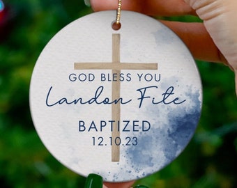 May include: A white round ceramic ornament with a blue watercolor background. A wooden cross is in the center with the text "God Bless You Landon Fife Baptized 12.10.23".