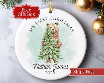 May include: A white ceramic Christmas ornament featuring a bear holding a Christmas tree. The ornament reads "My First Christmas" and includes the name "Nathan James" and the year 2025. The bear wears a sweater.