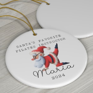K&ouml;nnte beinhalten: Wei&szlig;er Keramik-Weihnachtsbaumschmuck mit einer Cartoon-Abbildung des Weihnachtsmanns, der eine Pilates-Pose einnimmt. Der Text lautet "Santas Lieblings-Pilates-Lehrer Maria 2024".