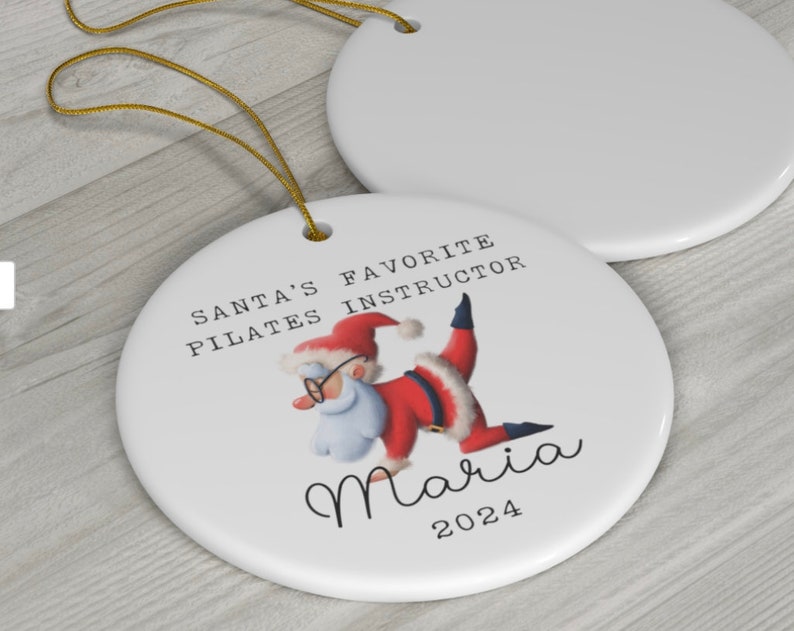 K&ouml;nnte beinhalten: Wei&szlig;er Keramik-Weihnachtsbaumschmuck mit einer Cartoon-Abbildung des Weihnachtsmanns, der eine Pilates-Pose einnimmt. Der Text lautet "Santas Lieblings-Pilates-Lehrer Maria 2024".