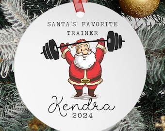 May include: White round Christmas ornament with a cartoon image of Santa Claus lifting a barbell. The text "Santa's Favorite Trainer" is above the image and the text "Kendra 2024" is below the image.