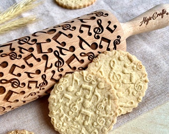 May include: A wooden rolling pin with a musical note pattern and the text "Algis Crafts" is next to several cookies with the same musical note design. The cookies are light brown and round. The rolling pin is light brown.