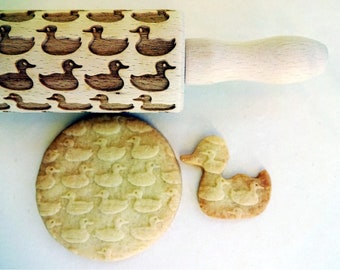 Puede incluir: Un rodillo de madera con un diseño de patos se muestra junto a dos galletas con forma de pato. Las galletas tienen el mismo diseño de patos que el rodillo. Las galletas son de color marrón claro y el rodillo es de madera clara.