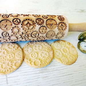 Könnte beinhalten: Ein Holz-Nudelholz mit einem geschnitzten Design aus Zahnrädern und Zifferblättern. Drei Kekse mit dem gleichen Zahnrad- und Zifferblattdesign sind unter dem Nudelholz zu sehen.
