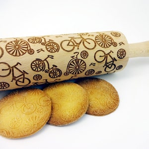Puede incluir: Un rodillo de madera con un patrón repetido de bicicletas y ruedas. El rodillo se utiliza para crear patrones decorativos en las galletas. Se muestran cuatro galletas con el patrón de la bicicleta en la imagen.