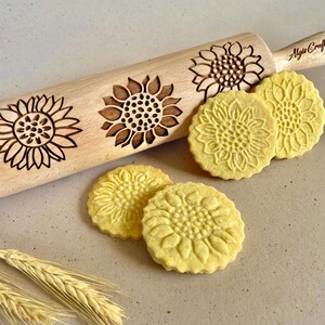 Può includere: Un mattarello di legno con quattro disegni di girasoli incisi sulla superficie. Quattro biscotti gialli con disegni di girasoli sono su una superficie beige accanto al mattarello.