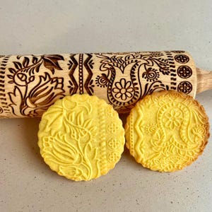 Könnte beinhalten: Ein Holz-Nudelholz mit einem komplizierten floralen und paisleyartigen Muster ist neben zwei Keksen mit dem gleichen Muster zu sehen. Die Kekse sind gelb und das Nudelholz ist braun.