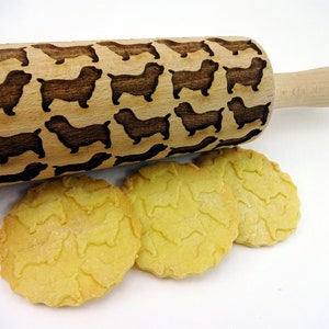 May include: A wooden rolling pin featuring a dachshund dog pattern, with three cookies displaying the same embossed design. The rolling pin has a natural wood finish, and the cookies are golden yellow.