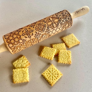 May include: A wooden rolling pin with a decorative pattern of geometric shapes and flowers. The rolling pin is engraved with the text "Algio Crafts". There are several cookies on a light gray surface, each with a different geometric pattern.