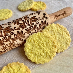 May include: A wooden rolling pin with a floral design engraved into the surface. The rolling pin is lying on a beige fabric surface with several yellow biscuits next to it. The rolling pin has the text "Algis Crafts" on the handle.