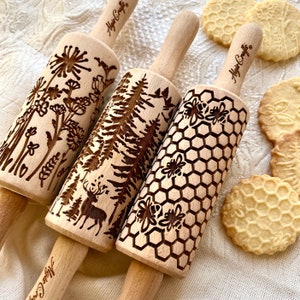 Puede incluir: Tres rodillos de madera con diseños grabados. Los diseños son una escena de bosque con un ciervo, un patrón de panal con abejas y un patrón floral con un diente de león. Los rodillos están sobre un fondo de tela blanca.