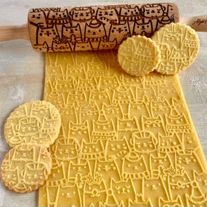 May include: A wooden rolling pin with a carved design of cats wearing hats and scarves. The rolling pin is used to create decorative patterns on dough. The image also shows cookies with the same cat design.