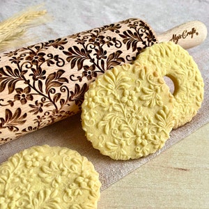 May include: A wooden rolling pin with a floral design engraved into the surface. Three cookies with the same floral design are next to the rolling pin. The cookies are light yellow and the rolling pin is brown. The rolling pin has the text "Algis Crafts" on the handle.