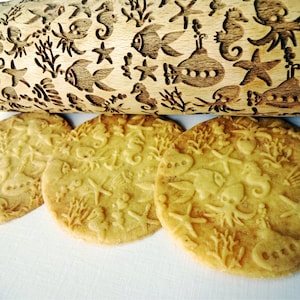 May include: A wooden rolling pin with an engraved underwater design featuring fish, seahorses, submarines, and starfish. Three round cookies with the same embossed pattern are in the foreground. The cookies are light brown.