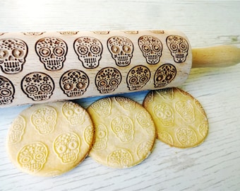 May include: A wooden rolling pin with a repeating pattern of sugar skull designs. Three round cookies with the same skull pattern are next to the rolling pin. The rolling pin has a light wood finish.