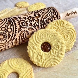 May include: A wooden rolling pin with a paisley design engraved into the surface. The rolling pin is used to create decorative patterns on cookies. Three cookies with the same paisley design are shown in the image. One cookie has a chocolate filling.
