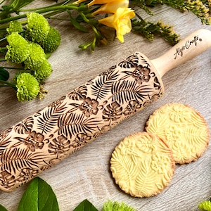 Pode incluir: Um rolo de madeira com um padrão de folhas e flores tropicais, juntamente com dois biscoitos com o mesmo design em relevo. O rolo tem uma pega de madeira clara e o texto "Algis Crafts". Os biscoitos são redondos e dourados.
