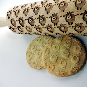 Op de afbeelding: Een houten deegroller met een gesneden ontwerp van koeien. Twee koekjes met hetzelfde koe-ontwerp zijn te zien onder de deegroller.
