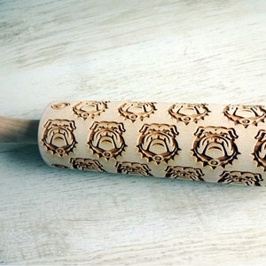 May include: A wooden rolling pin with a repeating pattern of bulldog faces engraved on the surface. The rolling pin is made of natural wood and has a light brown color.