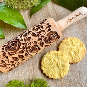 May include: A wooden rolling pin with a carved design of roses and leaves. The rolling pin is engraved with the text "Algis Crafts". Two cookies with the same design are shown next to the rolling pin.