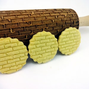 May include: Three round cookies with a brick pattern embossed on the surface. The cookies are light yellow and are sitting next to a wooden rolling pin with a brick pattern.
