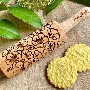May include: A wooden rolling pin with a floral pattern, alongside two cookies with the same embossed design. The rolling pin has the text "Algis Crafts" on the handle. The cookies are light yellow.