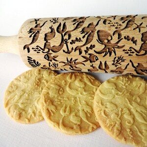 Pode incluir: Um rolo de massa de madeira com um design de sereia e criaturas marinhas. O rolo é usado para criar padrões decorativos em biscoitos. Três biscoitos com o mesmo design são mostrados abaixo do rolo.