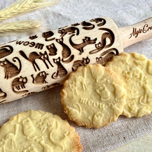 May include: A wooden rolling pin with a cat design. The rolling pin is engraved with cats and the words "meow" and "woom". The rolling pin is used to create cat-shaped cookies. The cookies are light brown and have a cat design.