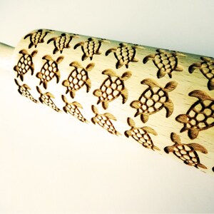May include: A wooden rolling pin with a repeating pattern of sea turtles carved into the surface. The turtles are detailed with a textured shell.