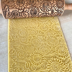Peut inclure: Un rouleau à pâtisserie en bois avec un motif de tournesol est utilisé pour imprimer un motif de tournesol sur une pâte. Le rouleau est brun clair et la pâte est jaune clair. Image en gros plan.