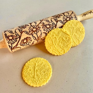 Puede incluir: Un rodillo de madera con un diseño floral de girasoles y hojas. El rodillo se utiliza para crear diseños en relieve en las galletas. Se muestran tres galletas con el mismo diseño de girasol junto al rodillo.