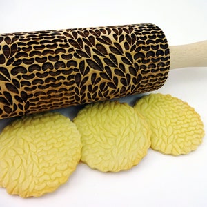 May include: A wooden rolling pin with a leaf pattern is displayed next to three cookies. The rolling pin has a light wood handle and a dark brown, engraved design. The cookies have a similar leaf pattern.