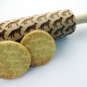 May include: A wooden rolling pin featuring a repeating pattern of dog silhouettes, with two round biscuits displaying the same embossed design. The rolling pin has a light wood handle and a patterned rolling surface. The biscuits are light brown.