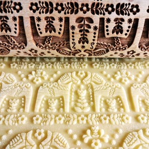 Puede incluir: Un rodillo de madera con un detallado patrón floral y de caballos se presiona en la masa, creando un diseño en relieve. El rodillo es de color marrón claro y la masa es de color amarillo pálido. El diseño incluye flores, hojas y caballos estilizados.