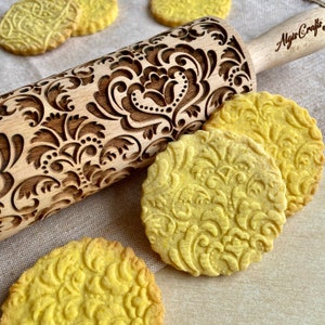 May include: A wooden rolling pin with an ornate floral pattern and the text "Algis Crafts" is displayed next to several round, embossed cookies. The cookies and rolling pin share a similar design, creating a cohesive baking theme.