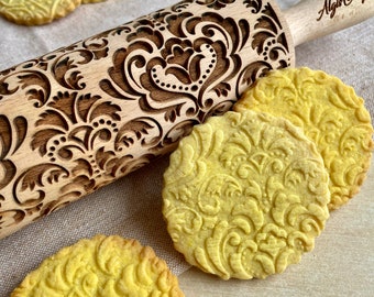 May include: A wooden rolling pin with an ornate floral pattern and the text "Algis Crafts" is displayed next to several round, embossed cookies. The cookies and rolling pin share a similar design, creating a cohesive baking theme.