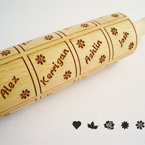K&ouml;nnte beinhalten: Ein Holz-Nudelholz mit vier personalisierten Namen: Alex, Kerrigan, Ashlin und Josh. Die Namen sind in einem quadratischen Muster mit einem Blumendesign in jeder Ecke graviert. Das Nudelholz hat einen Holzgriff.