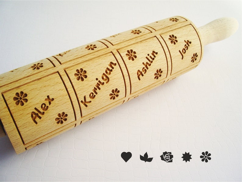 K&ouml;nnte beinhalten: Ein Holz-Nudelholz mit vier personalisierten Namen: Alex, Kerrigan, Ashlin und Josh. Die Namen sind in einem quadratischen Muster mit einem Blumendesign in jeder Ecke graviert. Das Nudelholz hat einen Holzgriff.
