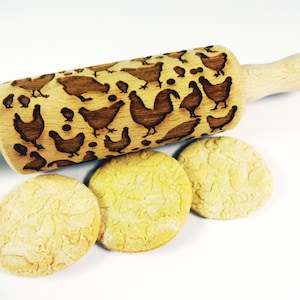 Op de afbeelding: Een houten deegroller met een ontwerp van kippen en kuikens. De deegroller wordt gebruikt om reliëfpatronen op koekjes of deeg te maken. Drie koekjes met het kippenontwerp zijn te zien onder de deegroller.