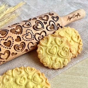 Puede incluir: Un rodillo de madera con un diseño de corazones y el texto "Algis Crafts" junto a tres galletas en forma de corazón. El rodillo es de color marrón claro y las galletas son amarillas. El rodillo mide aproximadamente 30 cm de largo.