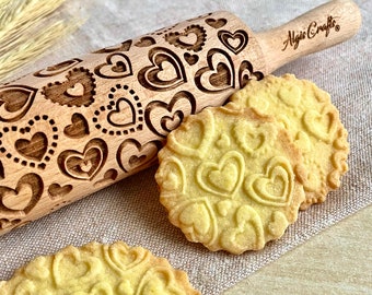 May include: A wooden rolling pin with a heart pattern and the text "Algis Crafts" is next to three heart-shaped cookies. The rolling pin is light brown, and the cookies are yellow. The rolling pin is approximately 12 inches long.