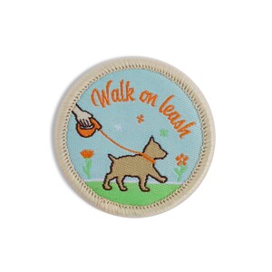 May include: A round, beige embroidered patch with a blue background. The patch features a cartoon dog on a leash being walked by a hand. The text "Walk on leash" is written in orange embroidery around the dog.
