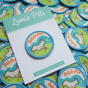 May include: A close-up of a blue and green embroidered patch with a white dog jumping in the air. The patch says "Fetch Pro" in orange text. The patch is surrounded by other similar patches, all with the same design. The text "Merit Badges For Dogs" is printed on a white card behind the patch.