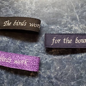 Puede incluir: Tres llaveros con texto bordado. Uno es negro con "The birds work", otro es morado con un estampado floral y "The birds work", y el tercero es azul marino con "for the bourgeoisie".