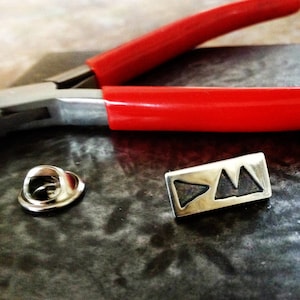 May include: A silver pin with a geometric design of three triangles. The pin is on a gray surface next to a pair of red pliers.