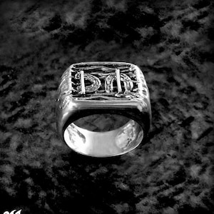 Puede incluir: Un anillo de plata con un diseño cuadrado que presenta intrincados patrones tejidos. El anillo es grueso y tiene un acabado pulido.