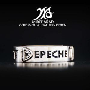 May include: Silver ring with the word "Depeche Mode" engraved on it. The ring is on a black surface.
